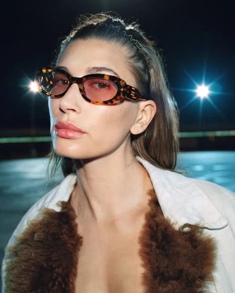 Hailey Bieber במשקפי שמש מאצטט בגוון צב עם עדשות ורודות, בלילה, בקמפיין סתיו-חורף 2026 של Miu Miu